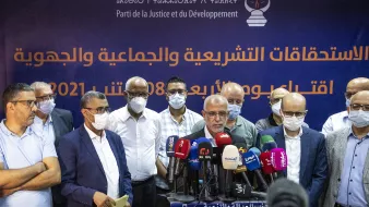تظهر في الصورة مجموعة من الرجال يقفون أمام لافتة تحمل شعار حزب العدالة والتنمية (Parti de la Justice et du Développement) في المغرب. يتحدث أحدهم، بينما يقف الآخرون خلفه، ومعظمهم يرتدون كمامات. يبدو أن هناك اهتمامًا كبيرًا من وسائل الإعلام، حيث تتواجد عدة ميكروفونات أمامهم. خلفهم، توجد جملة تشير إلى الاستحقاقات التشريعية والاجتماعية. المنظور العام يوحي بحدث رسمي يتعلق بالشؤون السياسية أو الانتخابات.