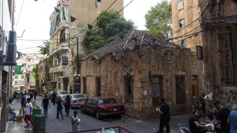 تظهر في الصورة زاوية من حي قديم في مدينة، حيث تتواجد مبانٍ قديمة ومتهالكة بجدران متصدعة. الشارع مزدحم ببعض الأشخاص الذين يسيرون ويقومون بأنشطة متنوعة. هناك سيارات متوقفة على جانب الطريق، وبعض الناس يجلسون في مجموعات. تتدلى بعض الزينة أو الأعلام من الأعلى، مما يضيف لمسة من الحيوية للمنطقة. الأشجار الخضراء تظهر في الخلفية، مما يُضفي طابعًا طبيعيًا على المشهد الحضري.
