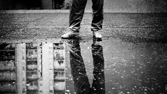 تظهر في هذه الصورة مجموعة من العناصر المثيرة للاهتمام. في المقدمة، نقف أمام شخص يرتدي بنطالًا و حذاء، واقفًا فوق سطح مغطى بالماء المتجمع، مما يشكل بركة صغيرة. يظهر انعكاس الشخص في الماء، مما يضيف بعدًا جماليًا للصورة. الخلفية عبارة عن جدار رمادي وسطح فارغ، مما يساعد على تركيز الانتباه على الشخص والانعكاس في الماء. الجو العام يبدو هادئًا ومظلمًا، مع تباين قوي بين الألوان.