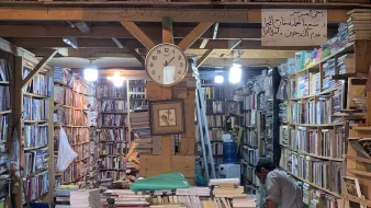 تظهر الصورة مشهداً داخلياً لمكتبة قديمة. تتراص الكتب على الرفوف الخشبية بشكل كثيف، مع ظهور بعض الكتب المكدسة على الأرض. في منتصف الصورة، يوجد ساعة حائط تشير إلى الوقت. كما يوجد شخص في الجهة اليمنى من الصورة، يبدو أنه يتفحص الكتب. الأضواء تتدلى من السقف، مما يضفي جواً دافئاً على المكان. في الأعلى، توجد لافتة مكتوبة توضح معاني تتعلق بالمكتبة.