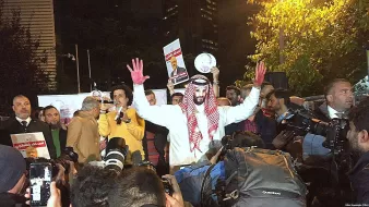 تظهر الصورة مجموعة من الأشخاص في حدث عام أو احتجاج. في الوسط، يوجد رجل يرتدي ملابس تقليدية وعليه علامة حمراء على يديه، مما قد يشير إلى رمز أو شعار معين. حوله، يوجد العديد من المصورين ووسائل الإعلام التي تسجّل الحدث. خلفية الصورة تحتوي على مبانٍ مرتفعة، مما يوحي بأن المكان قد يكون في مدينة كبيرة. يبدو أن هناك مزيج من الحماس والتوتر في الأجواء.