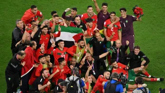 تظهر في الصورة مجموعة من اللاعبين والمشجعين المغاربة يحتفلون بشكل jubilant. يحمل اللاعبون العلم المغربي ويظهر عليهم الفرح والانتصار. يتمركز بعضهم حول مدربهم أو مدربين، بينما يتواجد آخرون بالقرب من كاميرات التصوير، مما يوحي بأجواء احتفالية قوية بعد تحقيق إنجاز رياضي. تعكس الصورة روح الفريق والوحدة بين اللاعبين.