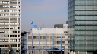 الصورة تظهر مبنى منظمة الدول المصدرة للنفط (أوبك) في وسط مدينة حديثة. يتكون المبنى من عدة طوابق ويتميز بتصميمه العصري. يوجد علم أوبك يرفرف فوق المبنى، كما أن هناك نوافذ كبيرة تسمح بدخول الضوء الطبيعي. المحيط يتضمن مبانٍ عالية أخرى، مما يشير إلى منطقة حضرية نشطة. الأجواء تبدو واضحة مع سماء زرقاء.