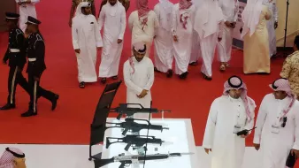 تظهر في الصورة مجموعة من الرجال العرب يرتدون الأزياء التقليدية، يتجولون في مكان يظهر فيه عرض للأسلحة. في وسط الصورة، توجد حاوية زجاجية تحتوي على عدة أنواع من الأسلحة، بما في ذلك البنادق. في الخلفية، يظهر بعض الأفراد المناسبين، مما يوحي بأن الحدث قد يكون معروضًا أو معرضًا للأسلحة أو الأمن. الأجواء تبدو رسمية وجادة، مع وجود السجادة الحمراء في الأرضية.