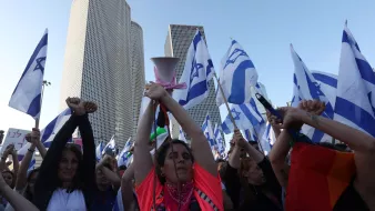 تظهر الصورة مجموعة من الأشخاص في مظاهرة، يحملون أعلامًا تحمل نجمة داود، وهو رمز معروف في إسرائيل. تظهر امرأة في المركز، مرفوعة اليدين وتحمل جهاز مكبر صوت، بينما يرفع الآخرون الأعلام. تتواجد خلفهم بنايات حديثة، مما يشير إلى أن المظاهرة قد تكون في منطقة حضرية. يعبّر المتظاهرون عن آرائهم، مما يدل على أهمية الفعالية التي يشاركون فيها.