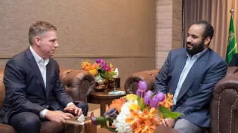 Two men in suits sit in a room, conversing near a floral arrangement on a table.