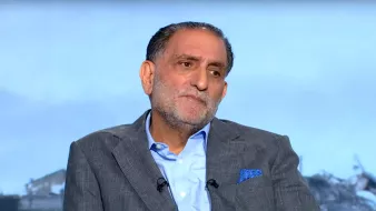 رجل يجلس مرتديًا بدلة رمادية، يبدو عليه التفكير والاهتمام.