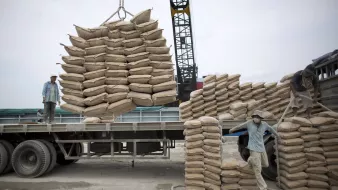 تظهر الصورة عمالًا يقومون بتحميل أكياس من مواد معينة على شاحنة. يبدو أن الأكياس مكدسة بعناية على الشاحنة، حيث يتم رفع بعضها باستخدام رافعة. يتركز العمل على تنظيم الأكياس وضمان تحميلها بشكل صحيح لتسهيل النقل. السماء تبدو غائمة، مما يوحي بأن العمل يتم في يوم غير مشمس.