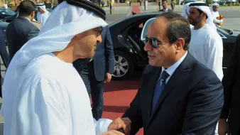 تظهر في الصورة شخصيات يرتديان ملابس تقليدية. الشخص على اليسار يرتدي الثوب الإماراتي الأبيض والغترة، بينما الشخص على اليمين يرتدي بدلة رسمية. يبدو أنهم يتصافحان في مناسبة رسمية، مع وجود سيارات فاخرة في الخلفية ومجموعة من الحراس أو الأشخاص الآخرين. تعكس الصورة أجواء من التعاون والاحترام المتبادل.