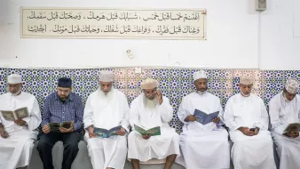 تظهر في الصورة مجموعة من الرجال يجلسون في صف واحد داخل مكان يبدوا أنه مسجد أو قاعة صلاة. كل منهم يحمل كتاباً، ويبدو أنهم يقرؤون أو يتدارسون. الجدران مزينة ببلاط ملون مزخرف، وهناك لوحة نصية مثبتة في الأعلى تحتوي على آيات أو نصوص دينية. يظهر في الصورة أجواء من التركيز والسكينة، مع تعبيرات الهدوء على وجوههم.