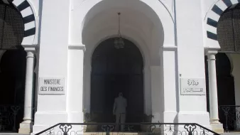 تظهر الصورة مدخل مبنى حكومي يُعرف باسم "وزارة المالية". يتميز المبنى بتصميم معماري جذاب، حيث يمكن رؤية الأبواب الكبيرة والنوافذ المزخرفة. المدخل محاط بأعمدة وأقواس، مما يعطيه طابعاً تقليدياً. هناك لافتات على الجانبين، واحدة باللغة الفرنسية والأخرى بالعربية، تشير إلى اسم الوزارة. يُلاحظ أيضاً شخص يقف أمام المدخل، مما يعكس النشاط في هذا المكان.