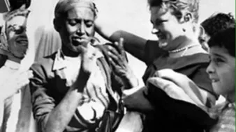 تظهر الصورة مجموعة من الأشخاص في احتفال يبدو أنه مليء بالحيوية والفرح. هناك شخص يظهر في المقدمة يرتدي ملابس تقليدية ويبدو أنه يشارك في عرض أو احتفال ثقافي. إلى جانبه، توجد سيدة تحمل ابتسامة على وجهها، مما يشير إلى جو من الألفة والود. الخلفية تحتوي على مزيد من الأشخاص الذين يظهرون في زوايا مختلفة، مما يعكس تفاعلًا اجتماعيًا نشيطًا. الألوان والتفاصيل تُسهم في الشعور بالبهجة والتفاهم بين الثقافات.