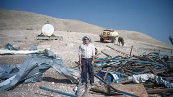 الصورة تظهر رجلًا standing في منطقة قاحلة، محاطًا بحطام وقطع من المعدن والمواد الأخرى. يبدو أن الرجل يرتدي ملابس تقليدية، وهناك خلفه مركبات ووحدات تخزين، كما تبدو الأرض غير مستوية وتنتشر فيها الصخور والحطام. السماء واضحة وزرقاء، مما يدل على يوم مشمس.