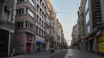 الصورة تظهر شارعًا في مدينة تبدو فيه الأجواء هادئة وخالية من الناس. تحيط بالمكان مبانٍ تاريخية بألوان دافئة، حيث تمثل العمارة الطراز المعماري المميز للمدينة. يبدو أن الشارع مُعدٌّ لوسائل النقل العامة، حيث توجد خطوط السكك الحديدية، ويمر ضوء خافت يعكس الصباح الباكر. في الوسط، يمكن رؤية كلب يسير عبر الشارع، مما يضيف لمسة من الحياة إلى المنظر الهادئ.