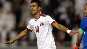 الصورة تظهر لاعب كرة قدم يرتدي زي المنتخب القطري، وهو يحمل الرقم 10. يبدو سعيدًا ويحتفل بعد تسجيل هدف أو تحقيق إنجاز في المباراة. خلفية الصورة تحتوي على جمهور أو لاعبين آخرين، مما يساهم في أجواء الحماس والفرحة الرياضية.