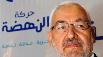 الصورة تظهر شخصًا يرتدي نظارات ويملك لحية وشعرًا أبيض، يبدو وكأنه يتحدث أو يعبر عن رأي. خلفه شعار يحمل اسم "حركة النهضة" والذي يشير إلى حزب سياسي في تونس. تظهر الألوان في الخلفية بشكل بسيط، مما يركز الانتباه على الشخص في المقدمة.