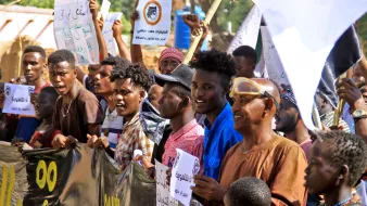 الصورة تظهر مجموعة من الناس في تظاهرة، يحملون لافتات و شعارات. يبدو أنهم متحمسون ويعبرون عن آرائهم. يظهر في الخلفية بعض المباني والأشجار، مما يدل على أن التجمع يحدث في منطقة مفتوحة. الوجوه تعكس مشاعر مختلفة، مثل الحماس والعزيمة.