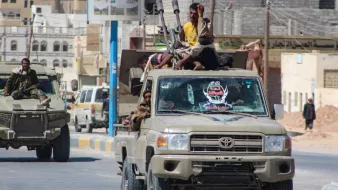 تظهر في الصورة مركبة عسكرية من طراز تويوتا، تحمل مجموعة من الرجال الذين يبدو أنهم في حالة استعداد. بعضهم يجلس على السقف، بينما يقودها شخص آخر في المقدمة. خلفهم، يمكن رؤية مبانٍ ذات طراز معماري مختلط، مما يوحي بأن الصورة قد التُقطت في منطقة حضرية. الجو العام مستقر إلى حد ما، على الرغم من وجود دلائل على التوتر. السيارة مزودة بأسلحة، مما يعكس سياقًا عسكريًا أو أمنيًا.