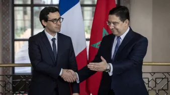 تظهر الصورة رجلين يرتديان بدلات رسمية. أحدهما يقف على اليمين ويمثل المغرب، حيث يظهر علم المغرب خلفه. الآخر يقف على اليسار ويمثل فرنسا، مع العلم الفرنسي خلفه. يبدو أن الرجلين يتصافحان في سياق رسمي، مما يشير إلى اجتماع أو تعاون بين البلدين. القاعة التي يتواجدان بها تبدو أنيقة وتحتوي على نوافذ كبيرة تسمح بدخول الضوء الطبيعي.