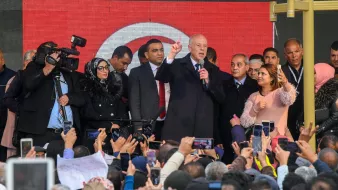 تظهر في الصورة حشود كبيرة من الناس تجمعوا في مكان عام. في المقدمة، يظهر شخص يقف على منصة ويتحدث، بينما يمسك العديد من الحضور هواتفهم المحمولة لتوثيق اللحظة. يحيط به مجموعة من الأشخاص، وبعضهم يبدو مشغولاً بالتقاط الصور أو الفيديو. خلف المنصة، توجد لافتة تحمل الألوان الوطنية. الأجواء توحي بالحماس والاهتمام من الحضور.