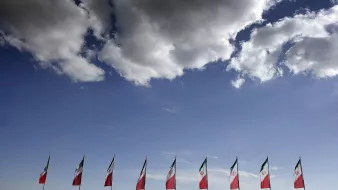 الصورة تُظهر مجموعة من الأعلام، حيث تكون الأعلام مرفوعة بشكل منتظم على طول الجزء السفلي. الأعلام تظهر بألوانها المميزة، مع خلفية من السماء الزرقاء التي تحتوي على بضعة غيوم بيضاء. يبدو أن الأعلام تمثل دولة معينة، والطابع العام للصورة يعطي شعورًا بالاحتفال أو الفخر الوطني.