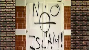 تظهر في الصورة بابًا مغطى بكتابات جدارية، حيث تم رسم عبارة "لا إسلام" مع رمز يظهر قوسًا دوائريًا. الكتابة تبدو باللون الأسود على خلفية بيضاء. الجدران المحيطة بالباب مصنوعة من الطوب بلون بني فاتح. الصورة تعكس مشاعر سلبية تجاه الإسلام، مما يثير جدلاً حول التسامح والقبول.