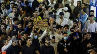 حشود من الناس في مكان مزدحم، يرفعون لافتة ويهتفون، مع تباين في الألوان والملابس.