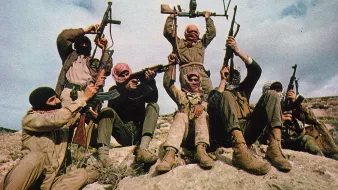 الصورة تظهر مجموعة من الأشخاص يرتدون ملابس عسكرية، وهم يتجهون نحو الكاميرا. بعضهم يحمل بنادق، بينما يقوم شخص في المنتصف برفع سلاح فوق رأسه. يظهر الأشخاص في وضعية متحمسة على صخرة كبيرة، وتظهر الخلفية سماء زرقاء مع بعض الغيوم. تعبّر الصورة عن روح الرفاقية والتصميم.