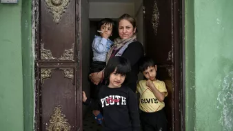 في الصورة، تظهر امرأة واقفة في مدخل منزل، تحمل سيجارة في يدها. خلفها، يوجد طفلان يلعبان، بينما يظهر طفل ثالث يقف أمامها. جدران المدخل ذات لون أخضر، والزخارف على الأبواب تظهر طابعًا قديمًا. تعكس تعابير الوجه في الصورة مزيجًا من الحذر والبراءة.