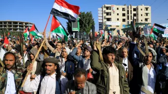 تظهر الصورة مجموعة من الأشخاص في تظاهرة حاشدة، يحملون الأعلام ويرتدون زيًّا تقليديًا. تعبر الوجوه عن الحماس وتظهر الأعلام التي تحمل ألوان الوطن. تتواجد خلفهم بنايات وبعض العلامات التجارية، مما يوحي بأن المكان هو منطقة حضرية. يبدو أن المتظاهرين يمارسون حقهم في التعبير عن آرائهم والمشاركة في حدث سياسي أو اجتماعي.