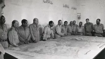 تظهر الصورة مجموعة من الرجال يرتدون زيًا عسكريًا، يجتمعون حول طاولة كبيرة تعرض خريطة. يبدو أن الاجتماع يدور حول التخطيط أو مناقشة استراتيجية ما. تعكس تعابير وجوههم التركيز والاهتمام بما يُناقش. الجدران خلفهم تبدو بسيطة، مع بعض الكتابات باللغة العربية. تُعطي الصورة انطباعًا عن موقف رسمي وعسكري.