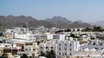 الصورة تُظهر مشهداً لمدينة محاطة بالجبال، حيث تتداخل المباني البيضاء مع بعض اللمسات الحديثة. تمتاز rooftops بأسقفها المسطحة، بينما تظهر في الخلفية الجبال الشاهقة التي تضفي جمالاً على المنظر. السماء نقية وزرقاء، مما يضيف إحساساً بالصفاء والهدوء للمنطقة.