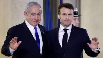 الصورة تظهر شخصين يقفان معًا أمام خلفية مميزة. يبدو أن أحدهما يرتدي بدلة زرقاء وقميص أبيض مع ربطة عنق زرقاء، بينما الآخر يرتدي بدلة سوداء وقميص أبيض مع ربطة عنق داكنة. كلاهما يبتسم قليلاً ويرفعان أيديهما في إشارة ودية. الجو العام في الصورة يبدو رسميًا، مما يشير إلى أن اللقاء قد يكون في مناسبة دبلوماسية أو رسمية.