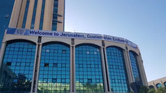 تظهر في الصورة مبنى يتميز بواجهته الزجاجية الواسعة. هناك لافتة كبيرة مكتوب عليها "مرحبًا بكم في القدس، سفارة غواتيمالا". يظهر في الخلفية سماء زرقاء وصافية، مما يعطي انطباعًا عن يوم مشمس. تنسيق المبنى يظهر حديثًا وعصريًا، مع بعض النباتات الخضراء في المقدمة.