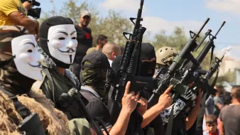 تظهر في الصورة مجموعة من الرجال يرتدون أقنعة وأزياء عسكرية، ويحمون أسلحة متنوعة. خلفهم يوجد عدد من الأشخاص في الخلفية. تعكس الصورة أجواء عسكرية أو استعراض لمجموعة مسلحة، حيث يتم التركيز على أسلحتهم وتنسيقهم. السماء تبدو زرقاء مع بعض السحاب، مما يعطي انطباعًا عن الأجواء المحيطة.