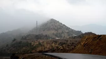 تظهر في الصورة مشهدًا جبليًا يكتنفه الضباب، حيث تتساقط السحب بشكل كثيف على الجبل. في الجزء العلوي من الجبل، يمكن رؤية برج اتصالات. الطريق يلتف حول الجبل، ويظهر بشكل مائل مع اختلافات في الألوان بين الصخور والتربة. الأجواء تبدو هادئة، مع لمسات من الغموض بسبب الضباب.