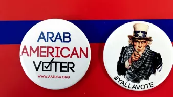 الصورة تحتوي على زرين دائرين. الزر الأول مكتوب عليه "ARAB AMERICAN VOTER" مع علامة التحقق، مما يدل على التصويت للمواطنين العرب الأمريكيين. الزر الثاني يظهر صورة لشخص يرتدي قبعة تحمل نجمة، وهو رمز معروف في الثقافة الأمريكية، مع عبارة "#YALLAVOTE" التي تدعو الناس للتصويت. الخلفية حمراء مع شريط أزرق، مما يضيف طابعاً احتفالياً للموضوع.