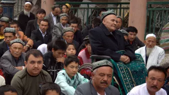 تظهر الصورة مجموعة من الأشخاص الجالسين في مكان مفتوح، يبدو أنهم مجتمعون في مناسبة أو تجمع. يوجد رجل مسن يجلس على كرسي متحرك في الخلف، ويبدو أنه يراقب ما يحدث من حوله. هناك تنوع في الأعمار بين الحضور، حيث توجد وجوه مراهقين وشباب وكبار في السن. يرتدي الكثير من الرجال قمصاناً تقليدية، وبعضهم يرتدي قُبُعات ذات تصاميم مميزة. يبدو أن الجو يسوده الانتباه والتركيز على الحدث الجاري.