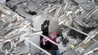 الصورة تظهر مشهداً من الدمار، حيث يوجد انخفاض في سقف أحد المنازل. debris متناثر ويرتفع الغبار في الجو، ويظهر شخصان يحاولان البحث عن شيء داخل المكان المدمر.