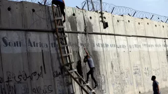 تظهر في الصورة جدارًا خرسانيًا عالياً يؤشر إلى وجود جهد للعبور أو تسلقه. يوجد على الجدار أسلاك شائكة في الأعلى، مما يضيف إلى الأجواء المشدودة. يتسلق أحد الأشخاص الجدار باستخدام سلم خشبي، بينما يحاول الآخر الصعود بشكل متكرر. الجدار مزين بخطوط ورسوم غريبة، بالإضافة إلى كتابات باللغة العربية. تعكس الصورة شعورًا بالجهد والمثابرة في مواجهة عوائق.