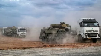 تظهر في الصورة مجموعة من الشاحنات تتحرك في منطقة صحراوية مغطاة بالتراب. تنتقل الشاحنات ببطء، وبعضها يحمل دبابات أو معدات عسكرية ثقيلة. يحيط بالمنطقة سماء ملبدة بالغيوم، مما يضفي جوًا من الحركة والنشاط إلى المشهد. يبدو أن هناك غبارًا كثيفًا يطير في الهواء نتيجة حركة الشاحنات، مما يخلق تأثيرًا دراميًا على الصورة.