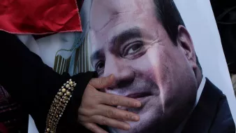 تظهر في الصورة يد تشير إلى صورة كبيرة لوجه شخص معروف، حيث توجد تعبيرات تدل على الارتياح والابتسامة. اليد تبدو وكأنها تعبر عن التأييد أو الولاء، بينما الخلفية تحتوي على ألوان تتضمن الأحمر، مما قد يشير إلى جو من الاحتفال أو التعبير عن الدعم.