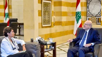 الصورة تظهر اجتماعًا داخل مكتب. يجلس رجل وامرأة على أريكتين. الرجل يبدو في منتصف العمر ويرتدي بدلة رسمية. المرأة ترتدي ملابس عادية، ويبدو أنها تشارك في الحوار. في الخلفية، تُرى ألوان العلم اللبناني، مما يدل على أن الاجتماع قد يكون ضمن إطار حكومي أو رسمي. الأثاث والتصميم الداخلي يعبّران عن الفخامة.