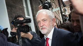 الصورة تظهر رجلًا متوسط العمر ذو لحية وشعر رمادي، يرتدي بدلة داكنة وربطة عنق حمراء. يبدو أنه في موقف يتطلب الانتباه، حيث تحيط به مجموعة من الكاميرات والصحفيين الذين يلتقطون صورًا له. الخلفية تعكس بيئة حضرية مع مبانٍ طوبية، وتعكس الحركة والاهتمام بالشخص.