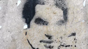 الصورة تظهر رسومًا جدارية تُظهر وجه إنسان بأسلوب الطلاء الرش. يبدو أن الرسم مقطوع بعض الشيء، مع تفاصيل غامضة لفم وأنف وعينين. الخلفية غير منتظمة وقد تشتمل على بعض آثار الندوب أو التآكل، مما يعطي الانطباع بأنها قد رسمت على سطح قديم أو متآكل. اللون المستخدم في الرسمة أسود بينما الخلفية ذات لون فاتح.