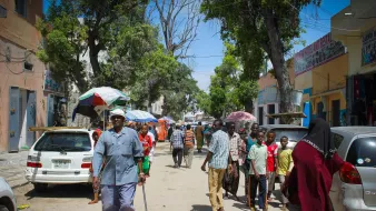 تظهر الصورة شارعا في منطقة حيوية، حيث يسير مجموعة من الأشخاص في اتجاهات مختلفة. في المقدمة، يوجد رجل مسن يستخدم عكازا للمشي، بينما يتواجد أشخاص آخرون في الخلفية، بعضهم يتحدث والبعض الآخر يسير. هناك سيارات متوقفة على الجانبين، وتظلل الأشجار والشمس المكان. يبدو أن الجو مشمس والناس يقومون بأعمالهم اليومية.