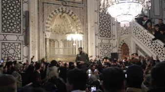 تظهر الصورة مجموعة من الأشخاص في مكان داخلي مزين بتفاصيل معمارية جميلة. هناك شخص يقف في الوسط، يبدو أنه يتحدث أو يلقي كلمة، بينما يحيط به حشد من المشاركين الذين يلتقطون الصور بأجهزتهم المحمولة. الإضاءة في المكان تتضمن ثريا كبيرة تعكس طابعًا تاريخيًا وثقافيًا، مع وجود عناصر زخرفية معمارية مميزة على الجدران. يبدو أن الحدث مهم وجذب انتباه الحضور بشكل كبير.