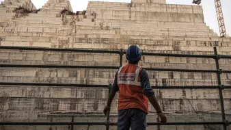 تظهر في الصورة عامل بناء يرتدي سترة واقية زاهية اللون وخوذة، وهو يقف أمام هيكل كبير من الخرسانة يبدو أنه في مرحلة الإنشاء. خلفه، تُلاحظ طبقات من المواد البنائية، مما يشير إلى مشروع بناء ضخم. يظهر في الخلفية معدات البناء مثل رافعة، مما يضيف إلى جو العمل المكثف والحركة في المكان. السماء تميل إلى اللون الأزرق، مما يوحي بأن الوقت هو إما الصباح الباكر أو بعد الظهر.