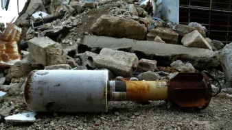 الصورة تظهر قنبلة أو جسم متفجر ملقى على الأرض، محاط بأحجار وأنقاض تشير إلى تدمير في المنطقة. يبدو أن الساحة غير مرتبة، والأرض مليئة بالردم والحطام، مما يشتت الانتباه عن الجسم المتفجر. يظهر أن هذا المكان شهد حدثًا مروعًا أدى إلى تدمير كبير.