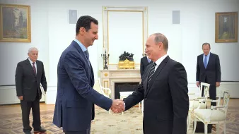 الصورة تظهر لقاءً بين شخصين رئيسيين، حيث يقوم أحدهما بمصافحة الآخر. الشخص على اليسار يرتدي بدلة زرقاء، بينما الشخص على اليمين يرتدي بدلة داكنة. يبدو أن اللقاء يجري في مكان رسمي، مع ديكور فخم بالإضافة إلى لوحات فنية على الجدران. في الخلفية، يوجد عدد من الأشخاص الآخرين الذين يشاهدون اللقاء بشكل غير مباشر. الجو العام يوحي بأهمية المناسبة.